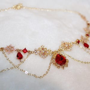 Könnte beinhalten: Ein goldfarbenes Kopfstück mit aufwendigen Details. Es verfügt über funkelnde, klare Kristalle und leuchtend rote Edelsteine in verschiedenen Formen, darunter Tropfen und Quadrate. Zarte Ketten hängen von dem Design, was ihm Eleganz verleiht.