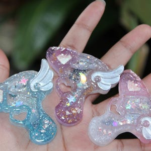 May include: Three resin gun-shaped charms in blue, purple, and pink, each with glitter and a wing design. The charms have a heart-shaped cutout and are filled with iridescent glitter and star-shaped accents.