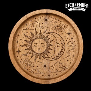 May include: A round wooden tray featuring a laser-engraved design of a sun and crescent moon with closed eyes, surrounded by stars, swirling galaxies, and clouds. The text Etch & Ember Studio is visible in the upper right corner of the image.