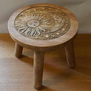 May include: A small, round wooden stool with three legs. The top is intricately carved with a sun and moon design, surrounded by stars. The wood has a natural, light brown finish, and the stool appears sturdy and decorative.