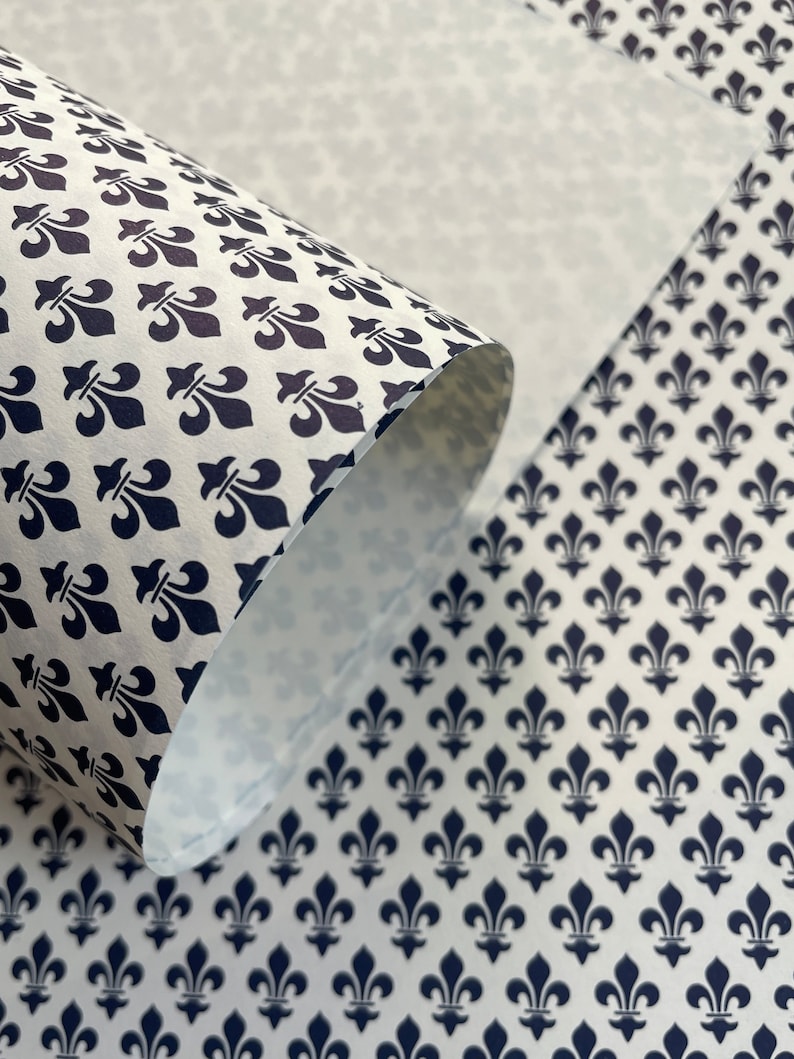 Puede incluir: Papel blanco con un patr&oacute;n repetitivo de flor de lis azul marino. El papel est&aacute; parcialmente enrollado, revelando el interior y la continuidad del patr&oacute;n. Adecuado para envolver, manualidades o fines decorativos.