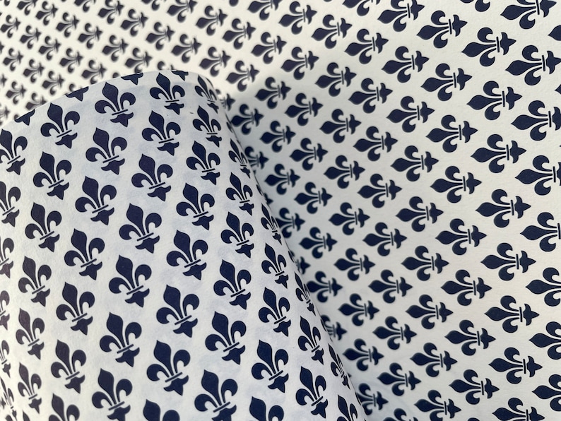 Puede incluir: Papel blanco con un patr&oacute;n repetido de s&iacute;mbolos de flor de lis azul marino. El papel est&aacute; parcialmente enrollado, revelando el patr&oacute;n en el interior. Adecuado para papel de regalo o proyectos de manualidades.