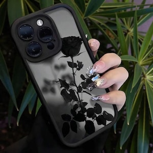 May include: Black phone case with a clear back and a black rose design. The case has a black border and a camera lens protector. The rose design includes the flower, stem, and leaves. The phone case is held in a hand with long, decorated nails.