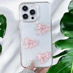 May include: A clear phone case with pink bow accents. The case is designed for an iPhone with a triple lens camera.