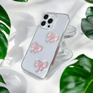 May include: A clear phone case with a white background and three pink bows. The case is designed for a smartphone with a camera bump.