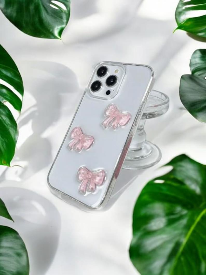 May include: A clear phone case with a white background and three pink bows. The case is designed for a smartphone with a camera bump.