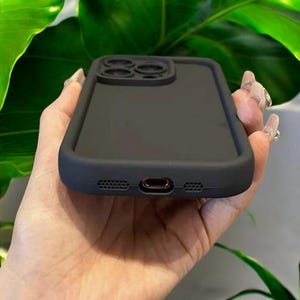 May include: A black silicone phone case with a raised camera lens protector. The case has a smooth, matte finish.