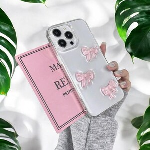 May include: A clear phone case with pink glitter bows. The case is designed for an iPhone with a triple lens camera.