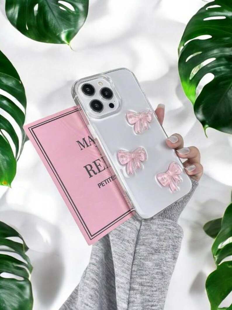 May include: A clear phone case with pink glitter bows. The case is designed for an iPhone with a triple lens camera.