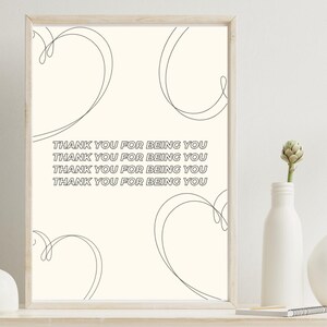 Peut inclure: Impression d'art encadrée avec un fond crème, avec le texte "THANK YOU FOR BEING YOU" répété quatre fois. Des cœurs en filigrane sont dessinés autour du texte. Un vase blanc avec une seule fleur est sur l'étagère.