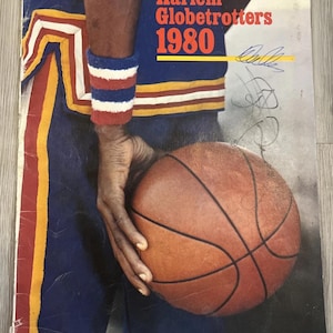 May include: Vintage Harlem Globetrotters 1980 program cover. The cover features a basketball player holding a basketball. The player is wearing a blue and yellow uniform with red and white accents. The program title is in red and yellow.
