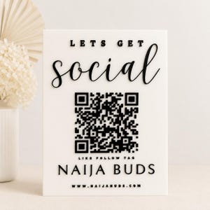 May include: A rectangular white sign featuring raised black lettering that reads LETS GET social, a central black QR code, the text LIKE FOLLOW TAG, the name NAIJA BUDS, and the website address www.naijabuds.com printed in a clear, sans-serif font.