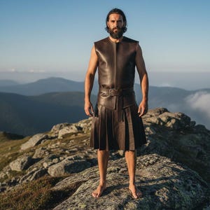 May include: A brown leather vest and kilt ensemble. The sleeveless vest has a zipper closure and the kilt is pleated with a belt. The outfit is worn outdoors on a rocky mountaintop with a mountain range in the background.