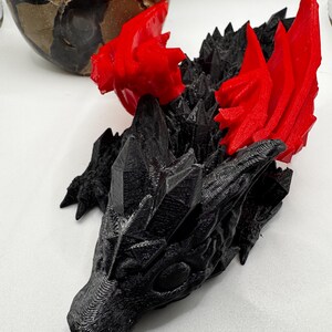 Könnte beinhalten: Ein 3D-gedruckter schwarzer Drache mit roten Flügeln. Der Drache liegt auf der Seite, der Kopf ruht auf dem Boden. Die Flügel sind hinter ihm ausgebreitet.