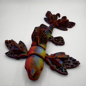 May include: A 3D printed, multi-colored, articulated insect sculpture with intricate wing details. The insect is red, orange, yellow, and blue.