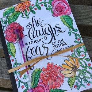 May include: A white journal with a black cover and a floral design. The journal is tied with a brown ribbon and has the text "She laughs without fear of the future. Proverbs 31:25" written on it.