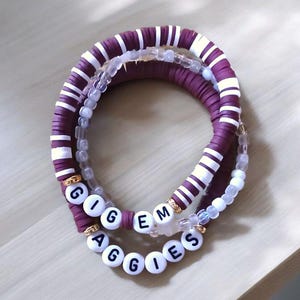 May include: Three stacked bracelets. The top bracelet is purple with white stripes. The middle bracelet is clear with white beads. The bottom bracelet has white letter beads spelling "GIG EM AGGIES".