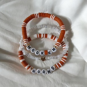 May include: Four stacked bracelets with a white background. The bracelets feature orange and white striped beads, white beads, and letter beads spelling "HOOKEM HORNS". One bracelet has a gold heart charm.