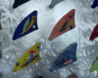 May include: A collection of colorful glass fin-shaped ornaments with abstract designs. The ornaments are arranged in a grid pattern on a silver background.