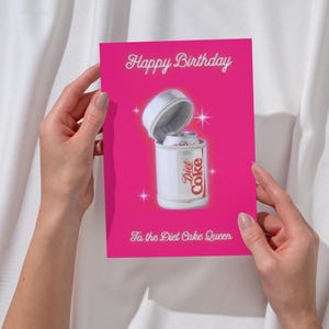 Pode incluir: Um cartão de aniversário rosa brilhante com a frase "Happy Birthday" em branco. O cartão apresenta uma lata de Diet Coke com a tampa aberta e o texto "To the Diet Coke Queen".