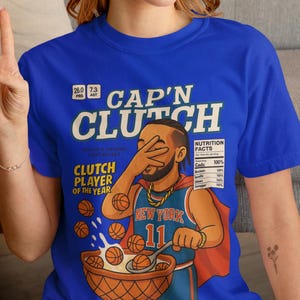Op de afbeelding: Een koningsblauw t-shirt met een grafische print van een basketbalspeler in een shirt met de tekst "NEW YORK 11" die basketballen uit een kom eet. De tekst "CAP'N CLUTCH" en "CLUTCH PLAYER OF THE YEAR" staat boven en naast de illustratie.