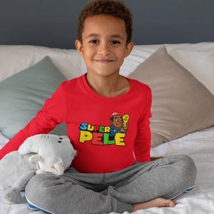 Peut inclure: Un t-shirt rouge à manches longues avec le texte "SUPER PELE" et un dessin animé. Le t-shirt est associé à un pantalon de survêtement gris. Un animal en peluche gris est au premier plan. La personne est assise sur un lit avec des draps et des oreillers blancs.