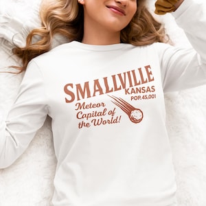 Op de afbeelding: Wit lange mouwen shirt met de tekst "Smallville Kansas" in bruin, samen met "Meteor Capital of the World!" en een afbeelding van een meteoor. Het shirt heeft een vintage design.