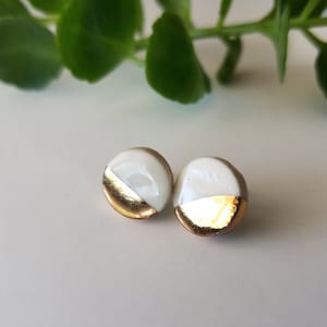 May include: A pair of small, round ceramic earrings with a white and gold design. The earrings are half white and half gold, with a sharp line dividing the two colors.
