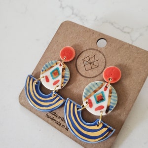 May include: A pair of handmade ceramic earrings with a blue and gold striped design, a white and blue geometric pattern, and red accents. The earrings are on a brown card with the text "andheartkc handmade in kc".