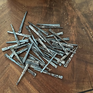 May include: A pile of weathered, rectangular metal nails with pointed ends. The nails are scattered on a dark brown wooden surface, showing signs of age and use. The nails are likely for woodworking or construction.