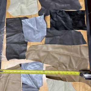 May include: Assorted leather scraps in various colors, including beige, gray, and black, are arranged on a wooden surface. A yellow measuring tape, showing measurements in inches, is placed across some of the leather pieces. The leather pieces have different textures.