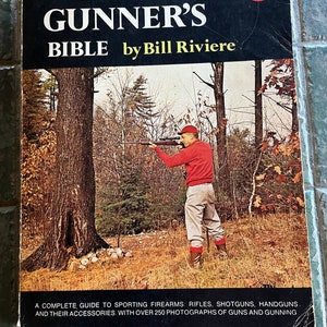 May include: A paperback book titled The Gunner's Bible by Bill Riviere. The cover features a person in a red sweater and cap aiming a rifle in a wooded area. The text reads: A complete guide to sporting firearms: rifles, shotguns, handguns.