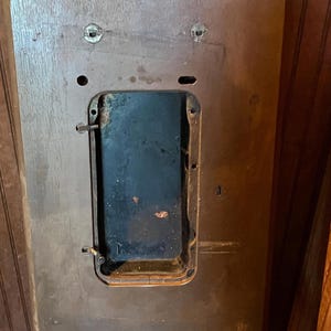 May include: A rectangular brown wooden sewing machine cabinet top featuring a central recessed black metal opening. The wooden surface displays a visible grain pattern, two circular mounting holes at the top, and a small metal latch mechanism on the left side of the central cutout.
