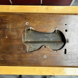 May include: A rectangular dark brown wooden sewing machine base with a central contoured cutout. The wood features a visible grain pattern and includes several metal mounting points, circular holes, and a recessed dark gray metal interior section.