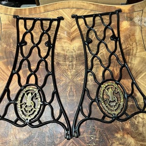 May include: Two black cast iron sewing machine side legs featuring an intricate lattice design. Each leg includes a central oval brass medallion embossed with the text THE SINGER MFG. CO. TRADE MARK and a central shuttle and needle graphic.