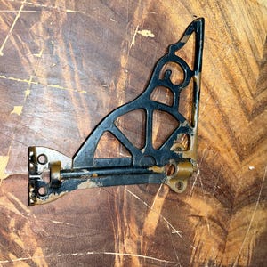 May include: A black metal shelf bracket with a triangular, openwork scroll design and worn gold-toned edges. The bracket features a horizontal bar with mounting holes at each end, showing signs of paint wear and metallic finish throughout the frame.