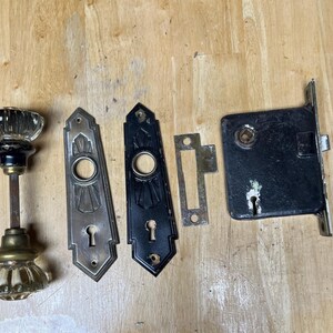 May include: A set of door hardware including a pair of clear glass knobs with brass and black accents on a metal spindle, two elongated metal backplates with keyholes, a small rectangular metal strike plate, and a black rectangular mortise lock.