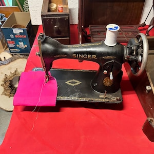 May include: A black vintage Singer sewing machine with gold decorative scrollwork, featuring the text The Singer Manufacturing Co. and SINGER. A white spool of thread sits on top, and a bright pink fabric scrap is positioned under the needle.