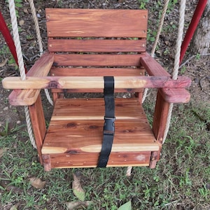 May include: A handcrafted wooden swing with a natural finish. The swing features a seat, backrest, and armrests, all made from reddish-brown wood. It includes a black safety strap and is suspended by thick, off-white ropes.