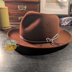 May include: A brown fedora hat with a wide brim and a ribbon band. The hat features a decorative feather and a small bow. The hat is sitting on a dark surface. The hat is a stylish accessory.