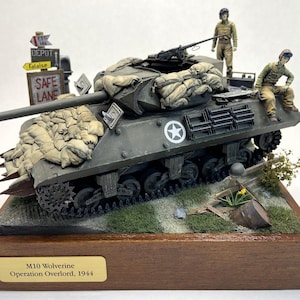 Puede incluir: Un modelo detallado de un tanque M10 Wolverine, pintado de verde con sacos de arena a los lados. Dos figuras con uniformes militares están de pie en el tanque. El tanque está sobre una base de madera con una placa que dice "M10 Wolverine, Operación Overlord, 1944".