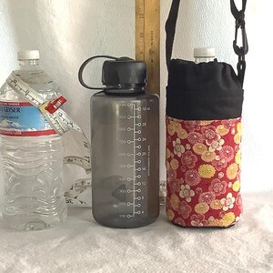 May include: A clear plastic water bottle with a label, a dark gray translucent water bottle with measurement markings, and a fabric water bottle carrier featuring a red floral pattern with a black top and an adjustable black shoulder strap.