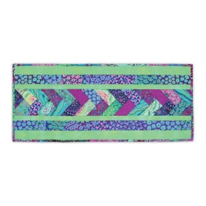 May include: A rectangular quilted table runner with a vibrant, colorful design. The runner features a central chevron pattern in shades of purple, teal, and green, framed by solid green borders and floral patterned strips. The overall aesthetic is bright and cheerful.