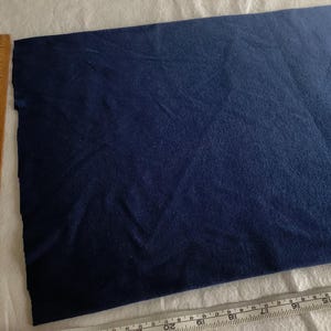 May include: A piece of navy blue fabric with a textured surface. A wooden ruler with inch markings is on the left side of the fabric. A clear plastic ruler with inch markings is at the bottom.