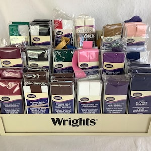 May include: A display of Wrights brand quilt binding and bias tape in various colors, including burgundy, black, white, and pink. The products are packaged in clear plastic and arranged in a tiered, off-white display case.