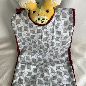 May include: A burgundy faux fur bib with a white cotton lining featuring a gray ginkgo leaf pattern. The bib has a rounded neckline.