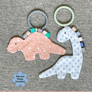 May include: Two fabric dinosaur sensory toys with silicone rings. One dinosaur is peach with white stars, the other is white with blue diamond patterns. The text "Dino Buddies" and "Sensory toys for baby to explore!" are visible.
