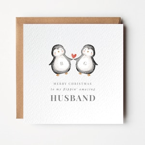 Puede incluir: Tarjeta de Navidad con dos pingüinos de acuarela sosteniendo un corazón rojo. La tarjeta dice "Merry Christmas to my flippin' amazing HUSBAND" en texto gris. La tarjeta tiene una superficie blanca texturizada y un sobre marrón.