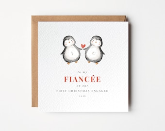 May include: A square white greeting card with a brown envelope featuring a watercolor illustration of two penguins holding a red heart. The text reads: to my FIANCÉE on our FIRST CHRISTMAS ENGAGED 2026. The penguins have the letters J and C on them.