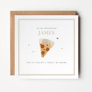 Può includere: Biglietto di auguri con l'illustrazione di una fetta di pizza. Il biglietto recita: "Al mio ragazzo James, hai rubato un pezzo del mio cuore". Il biglietto ha uno sfondo bianco con un bordo marrone ed è all'interno di una busta marrone.